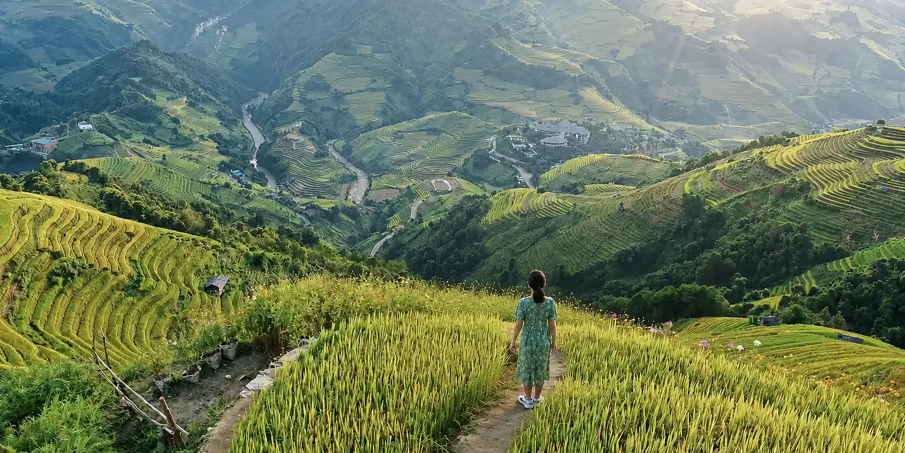 A traveler in a green dress walks a narrow path through golden rice terraces high above a misty Vietnamese mountain valley at sunrise